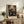 May include: Framed William Morris exhibition poster featuring a blue bird perched among vibrant orange and red flowers with decorative foliage against a cream background. The vintage-style botanical art print sits on a rustic wooden sideboard next to a fern plant, ceramic lamp, and books in a neutral-toned living room setting.