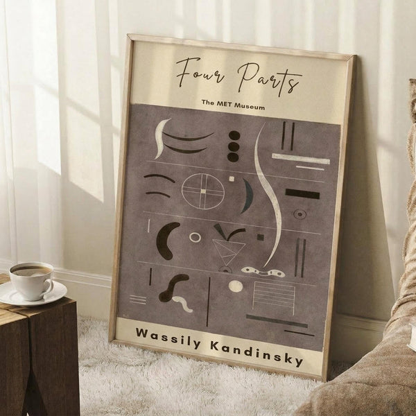 May include: Framed abstract art print leaning against white wall on textured cream rug. The poster features geometric shapes, lines and forms in muted brown and beige tones with museum-style text layout. A white coffee cup sits on a wooden block nearby, with a tan knitted throw visible at right. Natural window light creates soft shadows on the minimalist interior setting.