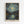 May include: Framed poster print of Van Gogh's Starry Night artwork mounted on white wall. Shows swirling night sky with bright yellow stars and moon over dark mountains and village with church spire. Water reflects lights from buildings and stars. Boat visible in foreground on right. Painting features thick, textured brushstrokes in blues, yellows, and dark tones. Natural wood frame with artist signature at bottom.