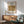 May include: Framed vintage William Morris angel artwork poster displayed above wooden sideboard in modern living room setting. Three robed figures in sepia tones within thin copper-colored frame. Mid-century green upholstered armchair with wooden arms, white ceramic table lamp, and round coffee table visible. Bright natural light creates shadows across white wall.