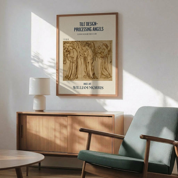 May include: Framed vintage William Morris angel artwork poster displayed above wooden sideboard in modern living room setting. Three robed figures in sepia tones within thin copper-colored frame. Mid-century green upholstered armchair with wooden arms, white ceramic table lamp, and round coffee table visible. Bright natural light creates shadows across white wall.