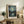 May include: Framed art print featuring swirling night sky with luminous stars and crescent moon over a lakeside town with mountain silhouettes, displayed on rustic wooden sideboard alongside fern plant, ceramic lamp, decorative vase, and stacked books in neutral-toned living room setting with beige upholstered chair and throw pillows