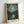 May include: Framed art print of Van Gogh's Starry Night painting featuring swirling blue-green night sky with bright yellow stars and crescent moon above dark mountains and village with reflective water. Print has artist attribution at bottom and sits in light wood frame against white wall, showing the classic post-impressionist brushstroke style with thick paint texture appearance.
