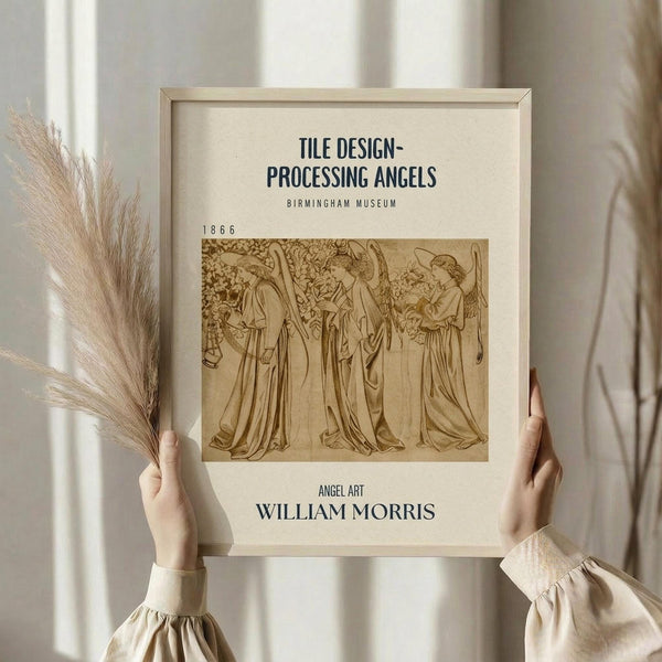 May include: Person in cream clothing holding framed vintage William Morris angel artwork from 1866 in light wooden frame. Print shows three robed angels in sepia tones on cream background with museum-style text layout. Dried pampas grass visible to left, soft natural lighting against neutral wall backdrop.