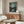 May include: Van Gogh's Starry Night poster print with swirling blue sky and golden stars, displayed in thin wood frame above walnut credenza. Modern living room setting with white curved chair, glass oval coffee table, and minimalist white table lamp. Soft natural lighting creates warm, contemporary atmosphere showcasing the classic impressionist artwork's vibrant blues and yellows against neutral beige walls.