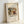 May include: A framed art print leaning against a white wall, featuring Wassily Kandinsky's abstract painting "Improvisation 27" from 1912. The artwork displays swirling organic forms in earthy tones of brown, yellow, green, and black with dynamic brushstrokes. The print is mounted on textured cream paper within a light wooden frame, creating natural shadows on the white surface below.