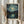 May include: Person in cream sweater holding framed poster print in light wood frame showing swirling night sky with bright yellow stars and crescent moon over dark village silhouette reflected in water, painted in thick impasto brushstrokes with blues, yellows and dark tones, displayed against white curtained window with dried pampas grass decoration