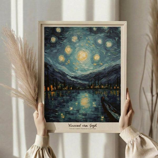 May include: Person in cream sweater holding framed poster print in light wood frame showing swirling night sky with bright yellow stars and crescent moon over dark village silhouette reflected in water, painted in thick impasto brushstrokes with blues, yellows and dark tones, displayed against white curtained window with dried pampas grass decoration