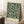 May include: Framed poster with dark green background featuring intricate cream-colored botanical pattern of flowing leaves, branches and small flowers in William Morris style, displayed leaning against white wall corner next to wooden side table with white coffee cup and beige knit throw blanket on textured rug.