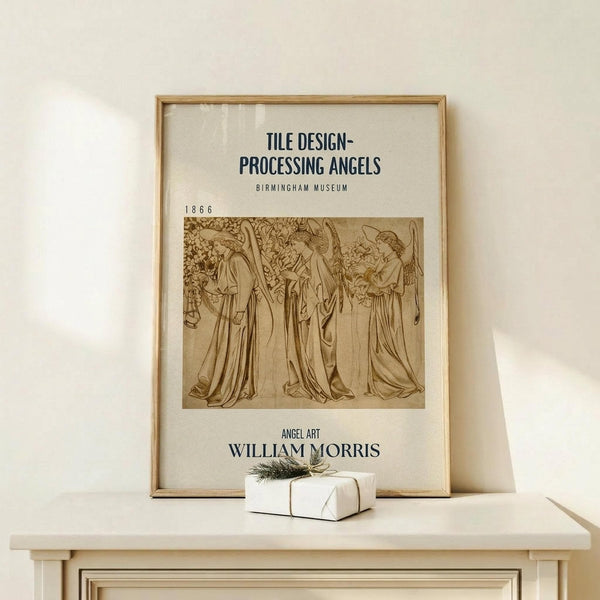 May include: Framed vintage-style poster featuring sepia-toned illustration of three angels in flowing robes from 1866, displayed on light wood shelf against cream wall. Small wrapped gift with greenery sits beside the natural wood frame. Museum exhibition poster format with text headers and William Morris attribution.
