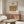 May include: Vintage William Morris angel artwork poster displayed above a mid-century modern walnut credenza in a minimalist living room. The beige and brown illustration shows three robed figures in medieval style. Scene includes white sculptural chair, glass oval coffee table, brass floor lamp, and modern table lamp with white shade.
