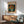 May include: Framed Van Gogh poster print of orange poppies in a pale green vase against a dark blue background, displayed above a mid-century modern wooden credenza with sliding doors. Scene shows mint green upholstered armchair with wooden frame, small round coffee table, and white ceramic table lamp. Warm natural lighting creates shadows across white walls in contemporary living room setting.