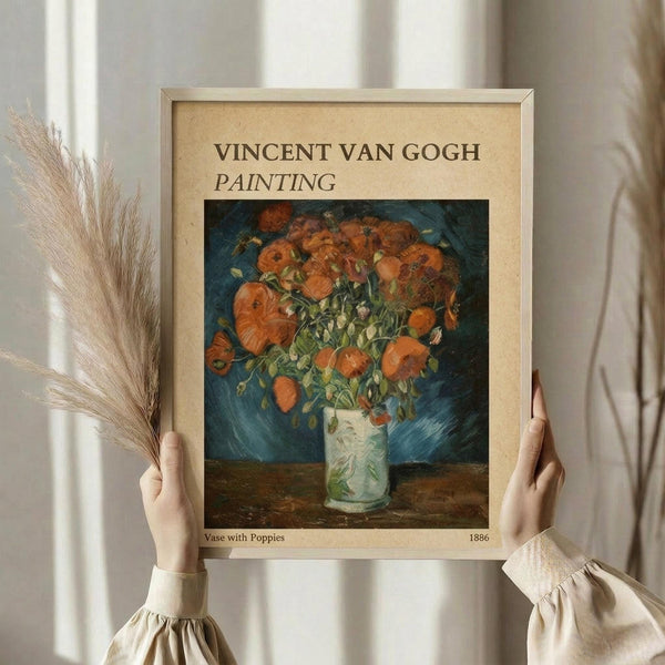 May include: Hands holding a framed reproduction of Van Gogh's vibrant orange poppies in a white vase against blue background, with dried wheat stalks visible to the left. The print features warm orange and red flowers with green stems in an impressionist style, displayed in a light wooden frame against white curtains and neutral clothing.