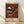 May include: Framed abstract art poster by Wassily Kandinsky titled "Gris" leaning against white wall on light wood floor. The artwork features geometric shapes including striped triangles, circles, and angular forms in rust orange, black, white, blue and gray tones against an orange background. Museum of New Zealand attribution appears at top. Natural lighting creates soft shadows across the scene.