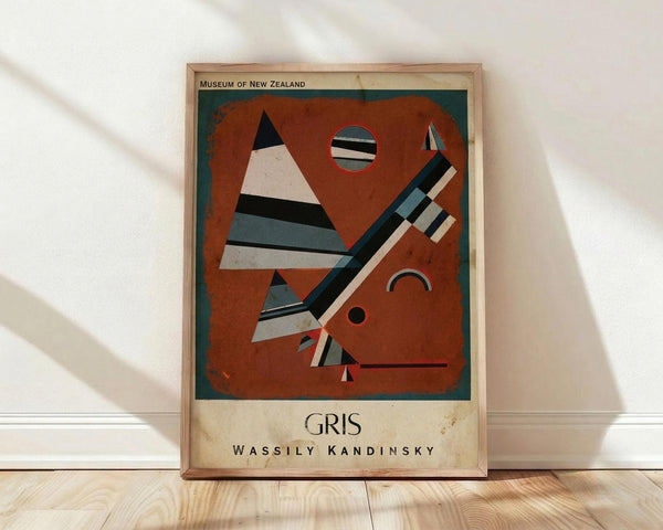 May include: Framed abstract art poster by Wassily Kandinsky titled "Gris" leaning against white wall on light wood floor. The artwork features geometric shapes including striped triangles, circles, and angular forms in rust orange, black, white, blue and gray tones against an orange background. Museum of New Zealand attribution appears at top. Natural lighting creates soft shadows across the scene.