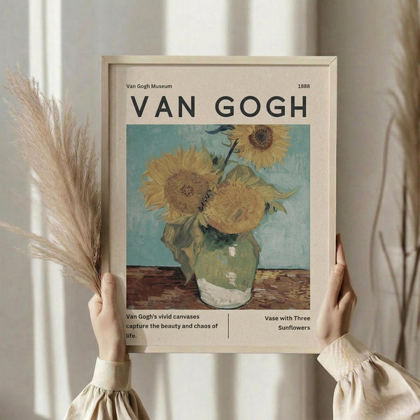 May include: Person in cream sweater holding framed Van Gogh museum poster featuring yellow sunflowers in green vase against light blue background. Poster has beige background with black text and museum-style layout. Dried wheat stems visible on left side. Soft natural lighting against white curtained window background.