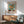 May include: Van Gogh sunflowers museum poster in light wood frame hanging on white wall above modern wooden sideboard. Green upholstered mid-century armchair with wooden arms sits nearby. White table lamp and round coffee table visible. Natural sunlight creates geometric shadows across the minimalist room setting.
