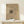 May include: Framed beige art print featuring abstract black linear drawing leaning against white wall on light wood surface. Small white wrapped gift with twine and greenery sits beside the thin wooden frame. Soft natural lighting creates gentle shadows on the minimalist display.