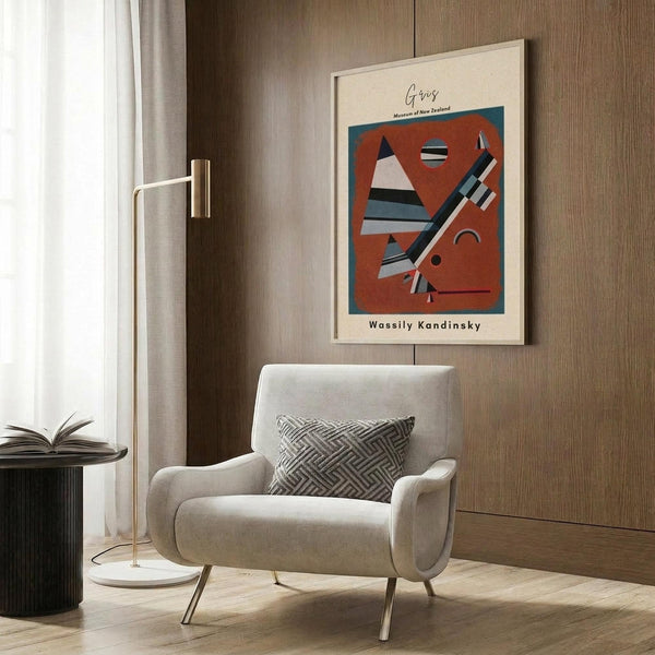 May include: Framed abstract art poster featuring geometric shapes in orange, white, and black on wood-paneled wall above cream upholstered armchair with geometric patterned pillow, brass floor lamp, and dark round side table with open book in modern living room setting.