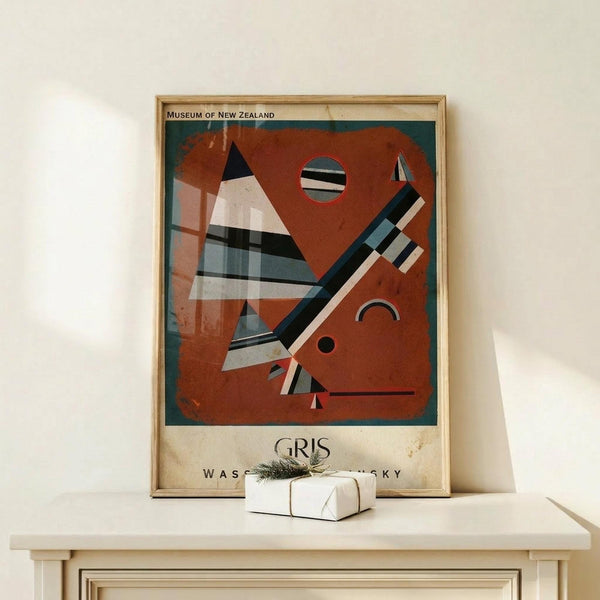 May include: Framed abstract art poster with geometric shapes in rust orange, teal, black and white stripes displayed on light wood surface. Features triangular and circular forms in modernist style. Small white gift box with twine sits beside the wooden-framed print against cream wall.
