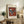 May include: Framed abstract art print with geometric shapes in orange, blue, and white tones leaning against white wall on rustic wooden sideboard. Scene shows cream armchair, fern plant, ceramic vase, stacked books, and warm natural lighting in modern living room setting.