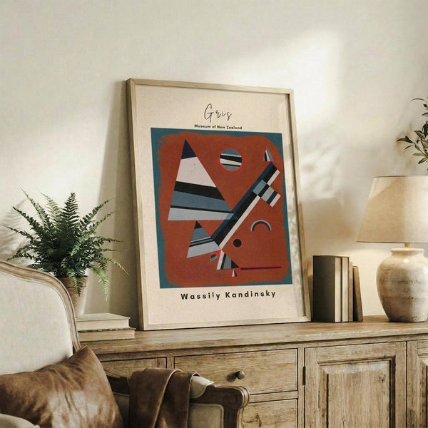 May include: Framed abstract art print with geometric shapes in orange, blue, and white tones leaning against white wall on rustic wooden sideboard. Scene shows cream armchair, fern plant, ceramic vase, stacked books, and warm natural lighting in modern living room setting.