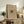May include: Beige framed art print displayed on rustic wooden sideboard in neutral living room setting. Abstract sketch artwork features dark flowing lines and organic forms on cream background. Frame sits among fern plant, stacked books, ceramic vase, and cream upholstered chair with brown throw pillows. Warm natural lighting creates soft shadows on white wall.