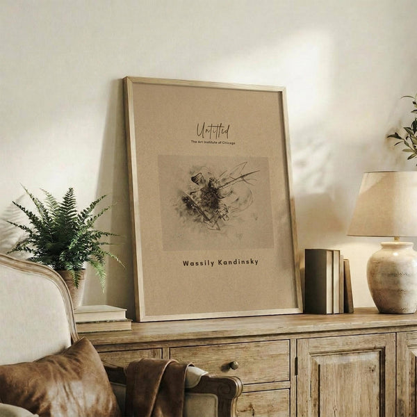 May include: Beige framed art print displayed on rustic wooden sideboard in neutral living room setting. Abstract sketch artwork features dark flowing lines and organic forms on cream background. Frame sits among fern plant, stacked books, ceramic vase, and cream upholstered chair with brown throw pillows. Warm natural lighting creates soft shadows on white wall.