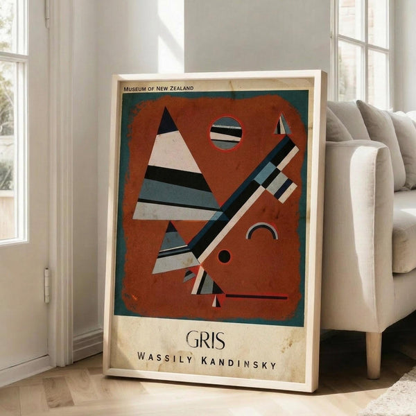 May include: Framed vintage-style museum poster featuring Wassily Kandinsky's abstract geometric artwork "Gris" with orange background, black triangular and striped shapes, and circular elements. The cream-colored poster shows "Museum of New Zealand" at top and artist name at bottom. Displayed leaning against white wall next to beige upholstered chair in bright, naturally-lit room with large windows and light wood floors.