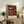 May include: Framed abstract art poster with geometric shapes in rust red, teal, black and white, displayed on rustic wooden sideboard alongside potted fern, ceramic vase, books and table lamp in warmly lit living room with cream upholstered furniture