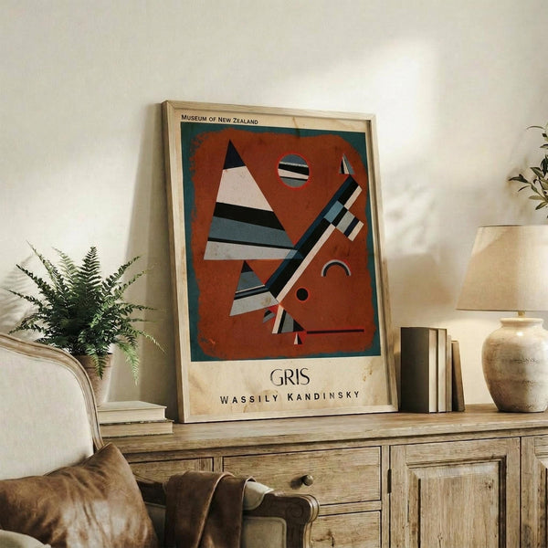 May include: Framed abstract art poster with geometric shapes in rust red, teal, black and white, displayed on rustic wooden sideboard alongside potted fern, ceramic vase, books and table lamp in warmly lit living room with cream upholstered furniture
