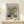 May include: Framed Van Gogh sunflower poster displayed on light wood surface against cream wall. The museum-style print features three yellow sunflowers in green vase against blue background, with exhibition text below. Natural wood frame, small wrapped gift beside it, plant shadows cast on wall creating serene home decor scene.