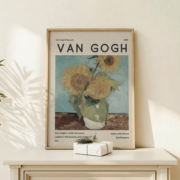 May include: Framed Van Gogh sunflower poster displayed on light wood surface against cream wall. The museum-style print features three yellow sunflowers in green vase against blue background, with exhibition text below. Natural wood frame, small wrapped gift beside it, plant shadows cast on wall creating serene home decor scene.