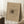 May include: A framed abstract art print with beige background featuring flowing black lines and organic shapes, displayed on a white textured rug in a bright room corner with natural light from windows creating geometric shadows on white walls, alongside a white coffee cup on saucer placed on a dark wooden block and neutral fabric.