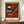 May include: Abstract modernist art print in wooden frame displayed on wooden sideboard next to white ceramic vase and stacked books. The artwork features geometric shapes in rust red, teal, black and white stripes arranged as triangles, circles and angular forms against an orange-red background. Museum-style poster with cream border and text at bottom. Wall has white wainscoting panels.