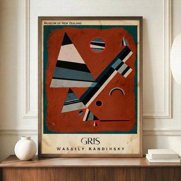 May include: Abstract modernist art print in wooden frame displayed on wooden sideboard next to white ceramic vase and stacked books. The artwork features geometric shapes in rust red, teal, black and white stripes arranged as triangles, circles and angular forms against an orange-red background. Museum-style poster with cream border and text at bottom. Wall has white wainscoting panels.