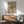 May include: Framed beige poster with abstract black ink drawing mounted on white wall above wooden sideboard. Mid-century modern living room setting with sage green upholstered armchair, round wooden coffee table, and ceramic table lamp. Natural sunlight creates diagonal shadows across the scene. The artwork features flowing, gestural black lines on cream background in thin wooden frame.