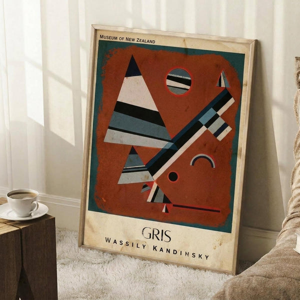 May include: Framed abstract art poster with rust-orange background featuring geometric shapes including striped triangles, circles, and angular forms in black, white, and blue tones. Text reads "Museum of New Zealand" at top and "GRIS Wassily Kandinsky" at bottom. Poster leans against white wall next to wooden block with white coffee cup and saucer, on textured cream rug with beige fabric visible.