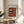 May include: Framed abstract art print in light wood frame displaying geometric shapes in orange, blue, black and white on cream background, positioned on round wooden side table next to beige upholstered chair with plant leaves visible in soft natural lighting.
