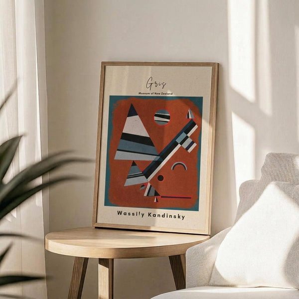 May include: Framed abstract art print in light wood frame displaying geometric shapes in orange, blue, black and white on cream background, positioned on round wooden side table next to beige upholstered chair with plant leaves visible in soft natural lighting.