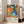 May include: Colorful abstract art poster in white frame leaning against white wall next to beige sofa. Artwork features vibrant floral and dot patterns in orange, blue, green, and pink with black and white polka dots. Text shows artist name and Paris museum reference with Japanese characters. Bright, modern living room setting with natural light from windows.