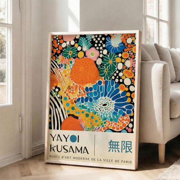 May include: Colorful abstract art poster in white frame leaning against white wall next to beige sofa. Artwork features vibrant floral and dot patterns in orange, blue, green, and pink with black and white polka dots. Text shows artist name and Paris museum reference with Japanese characters. Bright, modern living room setting with natural light from windows.