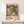May include: Framed vintage exhibition poster featuring vibrant abstract artwork with orange, blue, and green organic shapes, dots, and floral patterns on white background. Wooden frame leans against white wall on light wooden surface next to small wrapped gift with dried plant sprig.