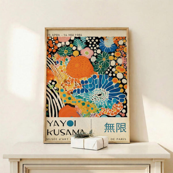May include: Framed vintage exhibition poster featuring vibrant abstract artwork with orange, blue, and green organic shapes, dots, and floral patterns on white background. Wooden frame leans against white wall on light wooden surface next to small wrapped gift with dried plant sprig.