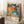 May include: Framed colorful abstract art poster leaning against white wall corner on fluffy white rug. Artwork features vibrant polka dots, floral patterns, and organic shapes in orange, blue, green, pink, and black with white text at bottom. Light wood frame, cozy interior setting with coffee cup on wooden block nearby.