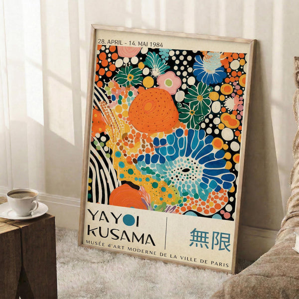 May include: Framed colorful abstract art poster leaning against white wall corner on fluffy white rug. Artwork features vibrant polka dots, floral patterns, and organic shapes in orange, blue, green, pink, and black with white text at bottom. Light wood frame, cozy interior setting with coffee cup on wooden block nearby.