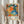 May include: Person holding framed colorful abstract art print with vibrant orange, blue, green polka dots and floral patterns. Text shows "YAYOI KUSAMA" and Japanese characters with Paris museum credit and 1984 exhibition dates. Light wooden frame against neutral curtain background with dried pampas grass visible to left side.