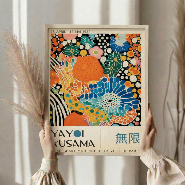 May include: Person holding framed colorful abstract art print with vibrant orange, blue, green polka dots and floral patterns. Text shows "YAYOI KUSAMA" and Japanese characters with Paris museum credit and 1984 exhibition dates. Light wooden frame against neutral curtain background with dried pampas grass visible to left side.