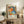 May include: Colorful abstract art poster with vibrant floral and geometric patterns in orange, blue, green, and pink tones, displayed in light wood frame on rustic wooden sideboard. Scene shows cozy living room setting with brown leather chair, potted fern plant, ceramic vases, and stacked books creating warm, modern home decor atmosphere.