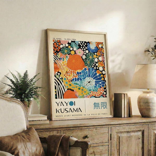 May include: Colorful abstract art poster with vibrant floral and geometric patterns in orange, blue, green, and pink tones, displayed in light wood frame on rustic wooden sideboard. Scene shows cozy living room setting with brown leather chair, potted fern plant, ceramic vases, and stacked books creating warm, modern home decor atmosphere.